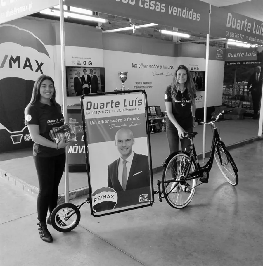 Stand da Remax num centro comercial com painéis impressos e uma bicicleta publicitária